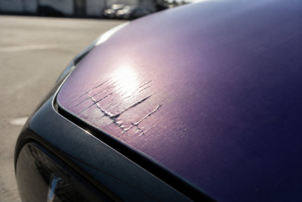 Close-up of a vehicle wrap surface showing visible color fading and slight cracking near the edge of the hood, with bright sunlight reflecting off the paint in the background