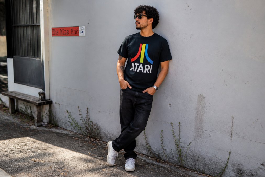 Person wearing vintage-inspired Atari graphic tee styled casually with dark jeans and white sneakers, standing in natural daylight with relaxed posture to show how retro prints integrate into everyday outfits; the print should be clearly visible at center-chest with intentional color separation