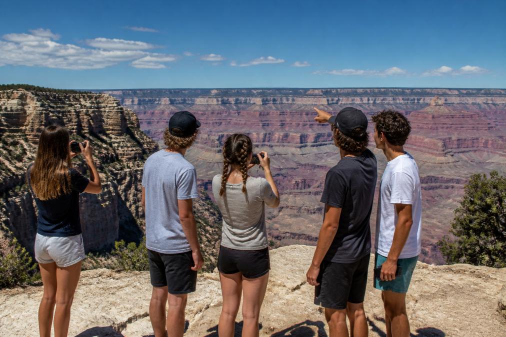 Group of teenagers standing at a scenic overlook at the South Rim, looking out across layered canyon walls with clear sky, some holding cameras or phones, appearing engaged and pointing at geological features
