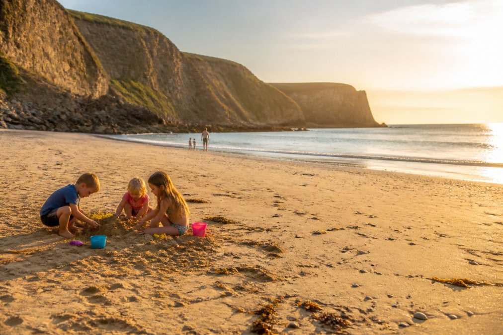 Family with children playing on sandy beach with dramatic sandstone cliffs in background, golden afternoon light reflecting off calm water