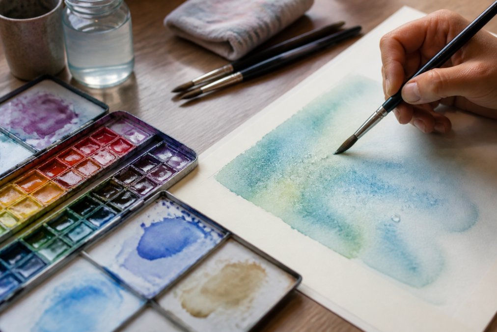 Close-up of watercolor palette with vibrant pigments, brushes, and a hand beginning a wet-on-wet wash on watercolor paper with water droplets visible
