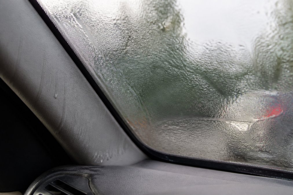 Close-up of water droplets and condensation on the interior of a car windshield, with visible moisture accumulated on the glass and trim, showing signs of interior water damage