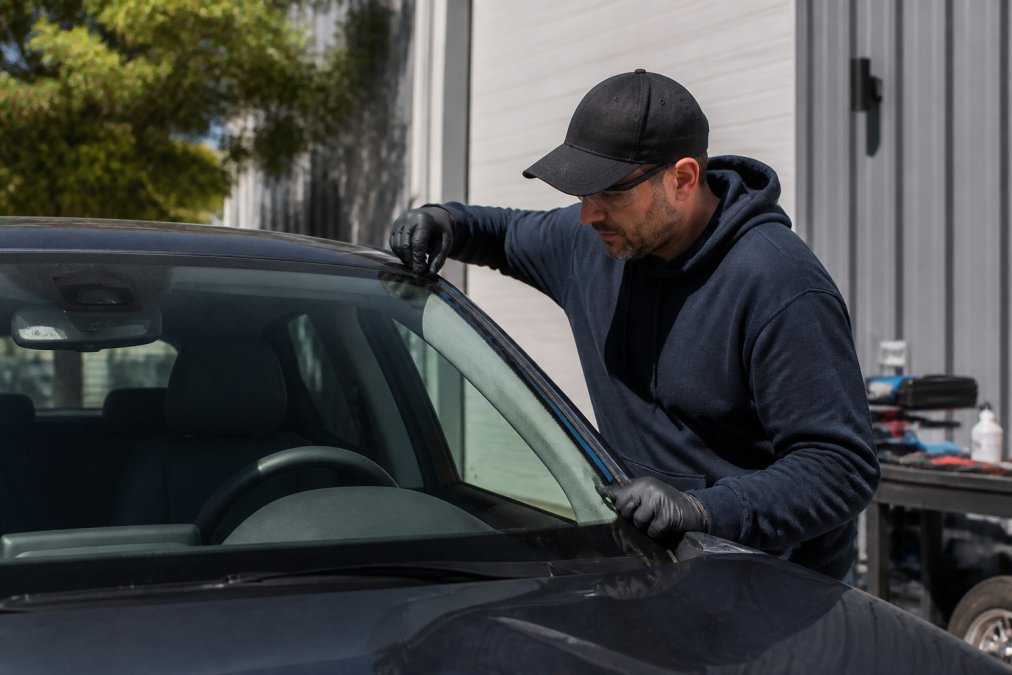 Professional auto glass technician in uniform carefully inspecting a freshly installed windshield, checking seals and alignment from multiple angles in natural daylight