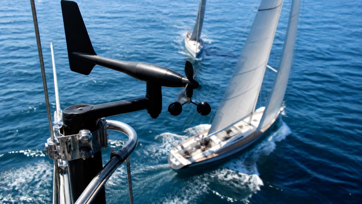 Close-up of a masthead wind transducer unit mounted on a racing sailboat mast with blue water and white sails visible below in bright daylight