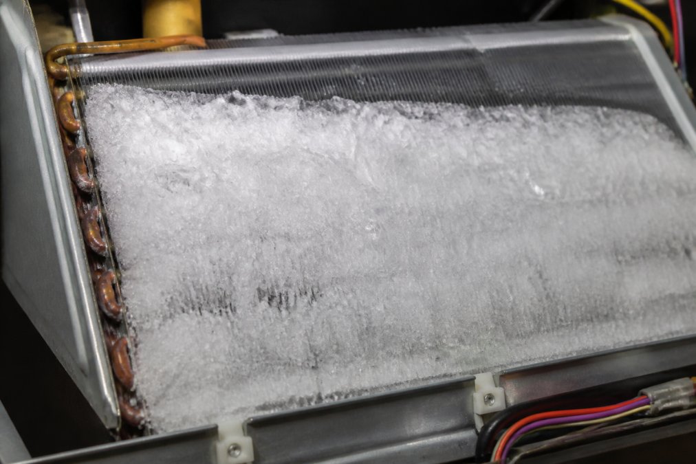 Close-up of a frozen evaporator coil covered in thick ice buildup, showing crystalline formations blocking the aluminum fins, photographed under bright workshop lighting to reveal the extent of the ice accumulation