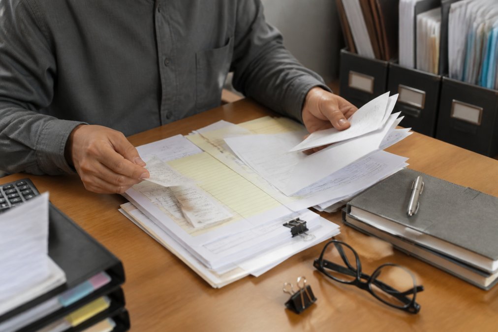 Close-up of hands organizing receipts, invoices, and financial documents on desk with checklist visible, organized filing system in background