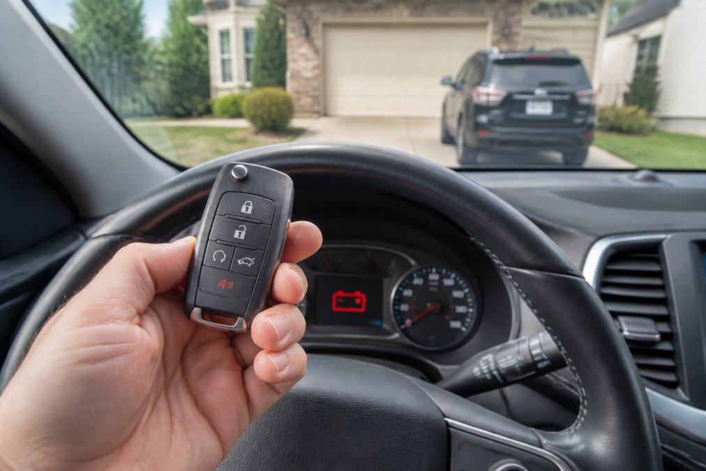 Close-up of a car key fob held in a person's hand, dead battery icon glowing on the dashboard behind it, parked in a suburban driveway in Northern Virginia
