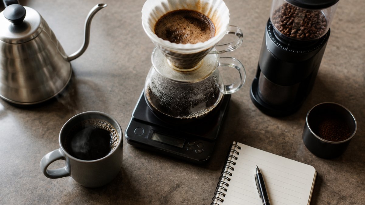 Overhead shot of a kitchen counter with a pour-over setup, digital scale, burr grinder, and a steaming mug of coffee, with a notebook and pen nearby for tracking adjustments
