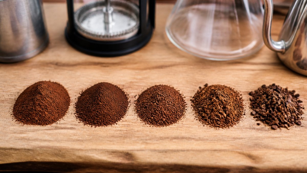 Close-up of coffee grounds in five distinct piles arranged left to right, showing progression from fine powder to coarse chunks, with brewing equipment partially visible in background on wooden surface