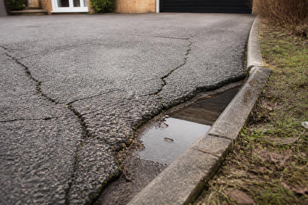 Close-up of a residential driveway showing visible cracks radiating outward, uneven surface with one side noticeably lower than the other, and water pooling in a sunken depression near the edge