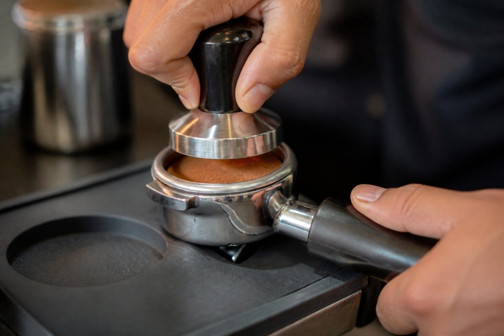 Close-up of a barista's hands tamping espresso grounds in a portafilter, showing proper tamping angle and pressure application over a coffee bed with even distribution