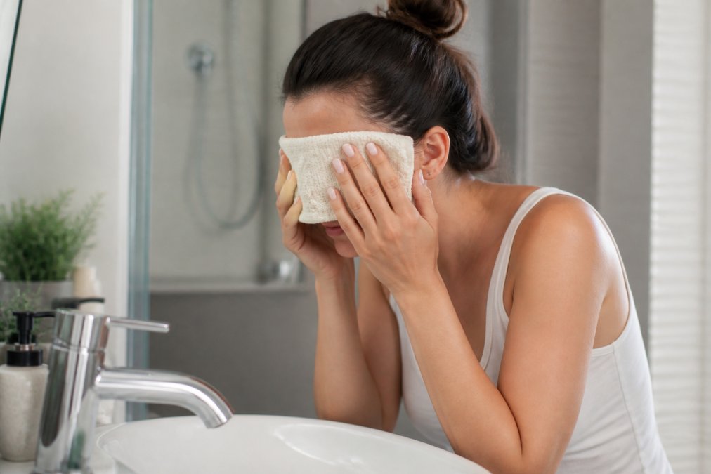 Close-up of hands holding a warm compress gently against the eyes over a sink in a bright bathroom, demonstrating the warm compress technique as part of a morning eye care routine