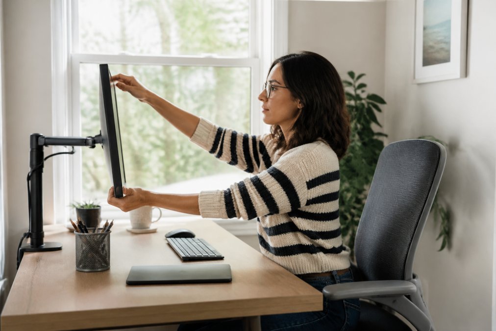 A person at a home desk adjusting the height and distance of a computer monitor, with a window providing soft natural light behind them