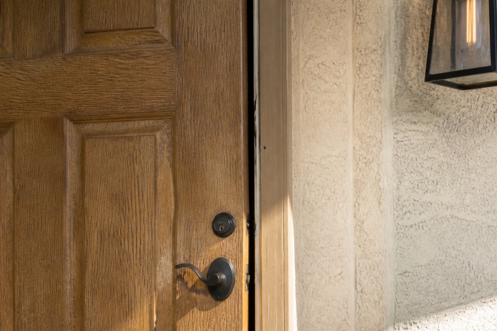 Close-up of a wooden front door showing visible bowing and separation gaps along the door jamb in bright afternoon sunlight