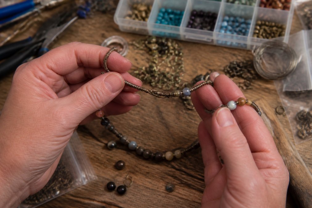 Close-up of hands examining a handmade beaded necklace with visible tarnished metal findings and uneven wire wrapping on a wooden workbench