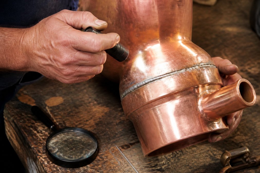 Distiller inspecting a copper still joint for metallic moonshine quality using a flashlight and magnifying glass