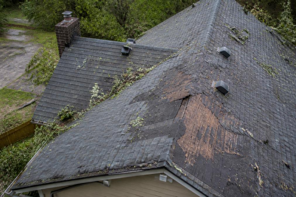 Aerial view of a residential roof showing missing shingles, debris scattered across the surface, and visible dark water damage stains on the roof deck after a severe storm