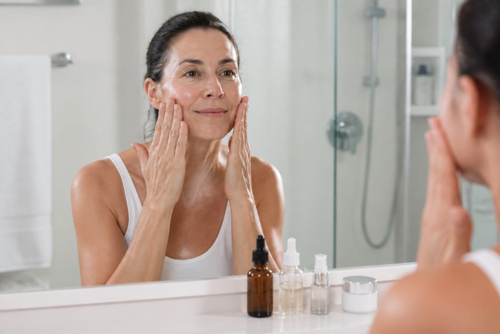 Woman in her late 40s gently pressing tight skin serum into damp cheeks in a bright bathroom setting