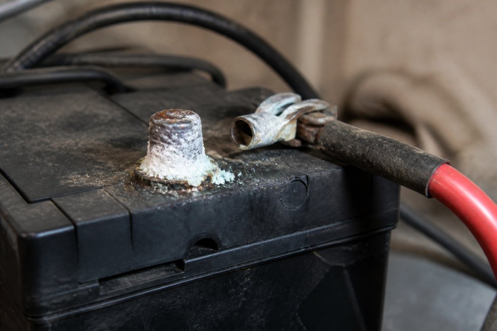 Close-up of a sump pump battery terminal showing white corrosion buildup and a loose cable connection in a basement environment with dim lighting