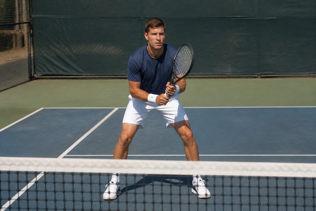 Tennis player at the net demonstrating proper ready position with feet shoulder-width apart, knees slightly bent, racket up at shoulder height, showing split-step positioning with natural court lighting