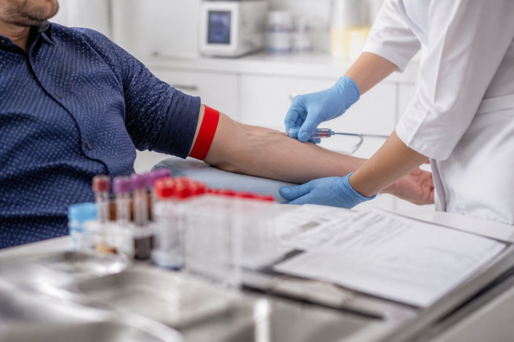 Laboratory technician drawing blood from a patient's arm in a clinical setting, with a requisition form showing testosterone test parameters visible on the desk, modern lab equipment in the background