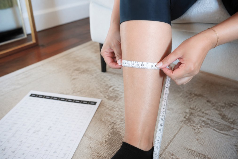 Woman's leg with measuring tape wrapped around the widest point of the calf, showing proper measurement technique with a size chart visible on a white surface nearby