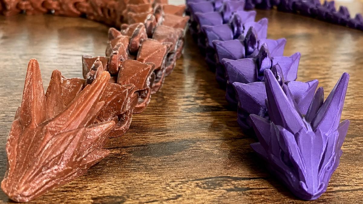 A close-up photograph of two articulated 3D printed dragon figures side by side on a wooden desk, one with visible joint wear and one with tight, clean joints, natural lighting highlighting the texture differences