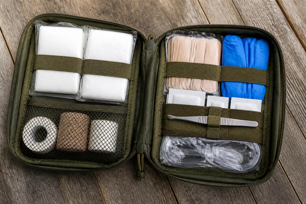 Flat lay of an open wilderness first aid kit on a wooden surface showing sterile gauze pads, adhesive bandages, nitrile gloves, antiseptic wipes, medical tape, tweezers, and a CPR face shield neatly arranged in labeled compartments under natural daylight
