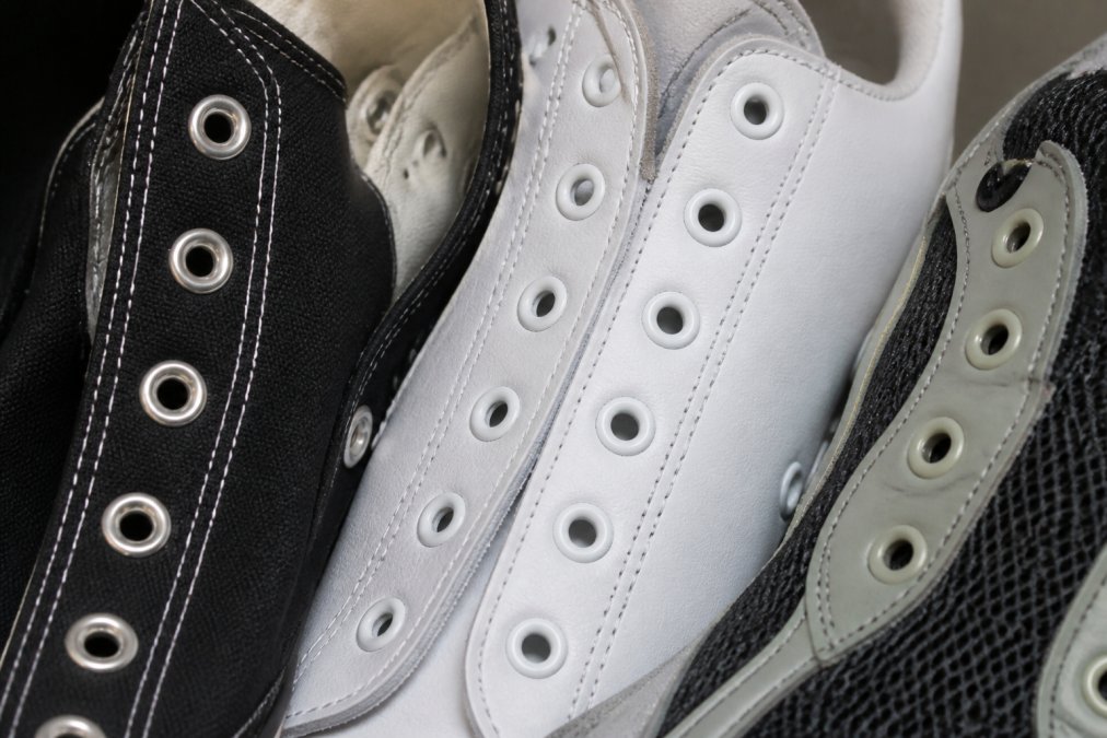 Close-up of various sneaker eyelets showing different spacing patterns, hole sizes, and materials including metal grommets on canvas shoes, reinforced eyelets on leather sneakers, and mesh eyelet trim on athletic shoes under natural lighting
