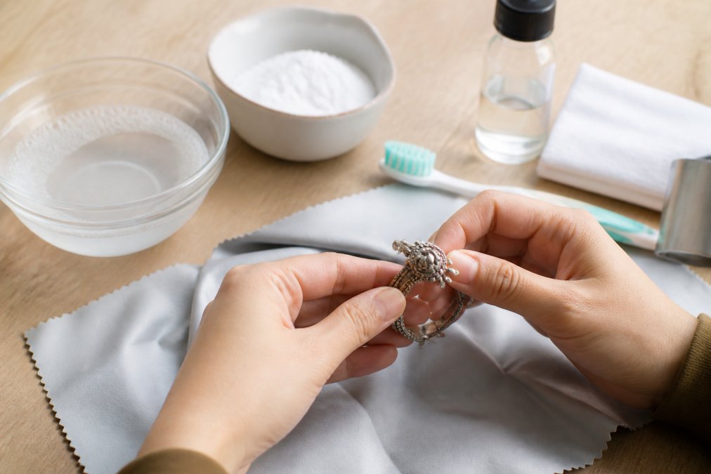 Hands holding a piece of tarnished sterling silver jewelry over a soft polishing cloth, with cleaning supplies arranged on a light wooden surface in natural daylight