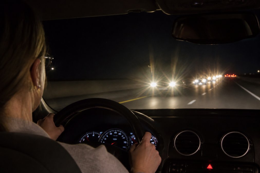 A person driving at night on a highway, squinting slightly at oncoming headlights that appear with visible halos and starbursts around them, dashboard softly lit