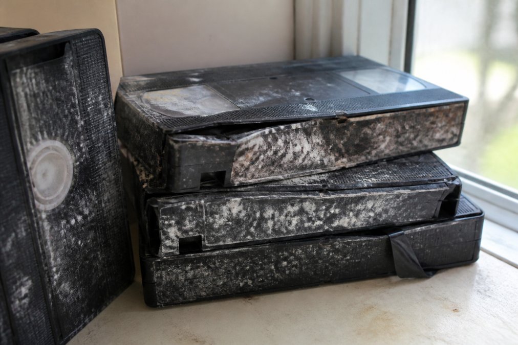Close-up of deteriorating VHS tapes showing visible white and brown mould spots, discoloured sections, warped plastic casing, and brittle tape edges on a shelf with natural window lighting
