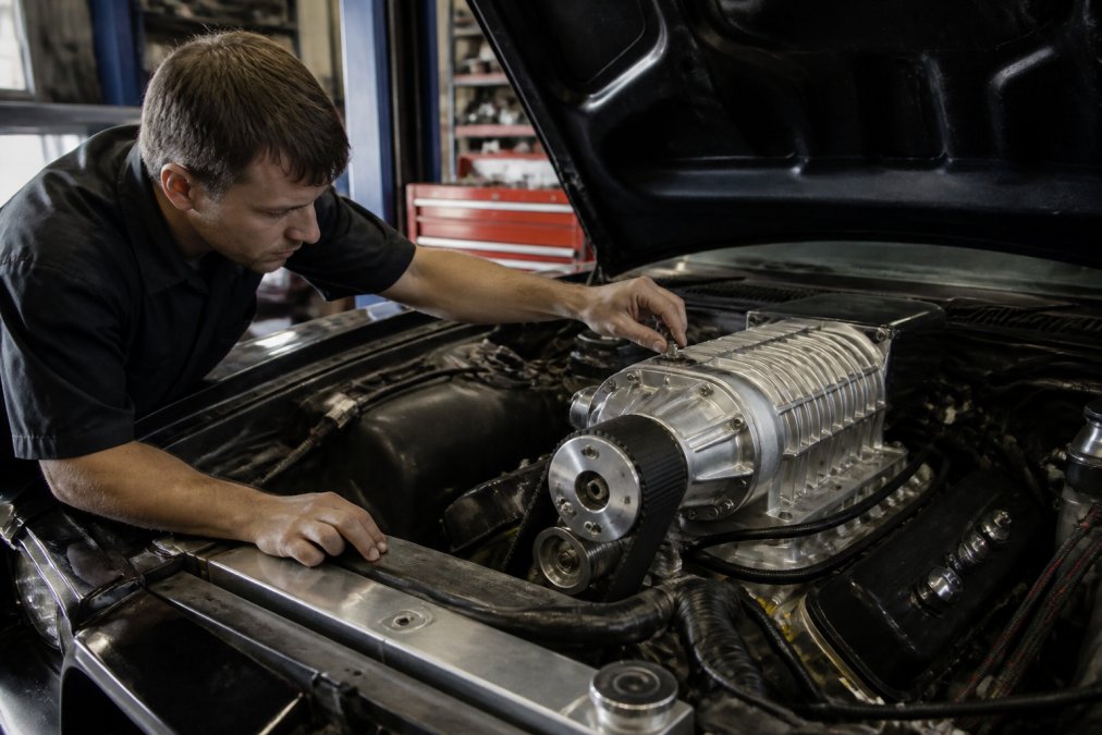 Professional mechanic examining a belt-driven supercharger installed on a V8 engine in a well-lit workshop, with visible intake manifold and polished aluminum components