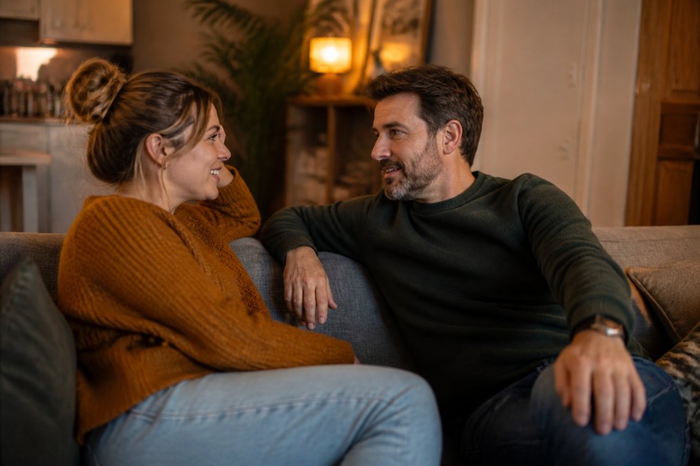 Een koppel zit samen op een bank in een warm verlicht huis, in gesprek met elkaar, met oogcontact en ontspannen lichaamstaal