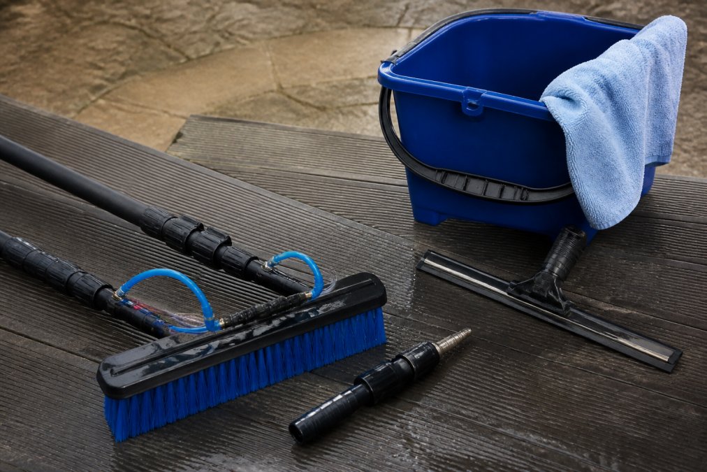 Close-up of professional window cleaning tools arranged on work surface including squeegee with replaceable rubber blade, microfibre cloths, bucket with handle, and water-fed pole components in natural workshop lighting