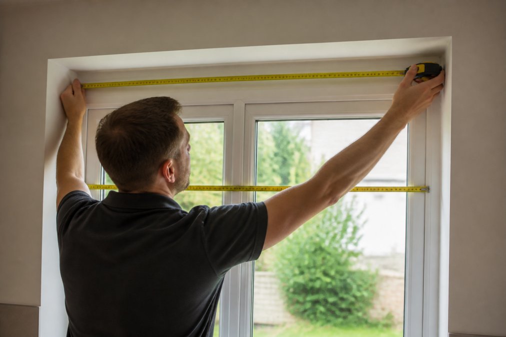 Professional demonstrating precise window measurement technique using tape measure against window frame at multiple points, showing recess fit positioning with natural daylight streaming through
