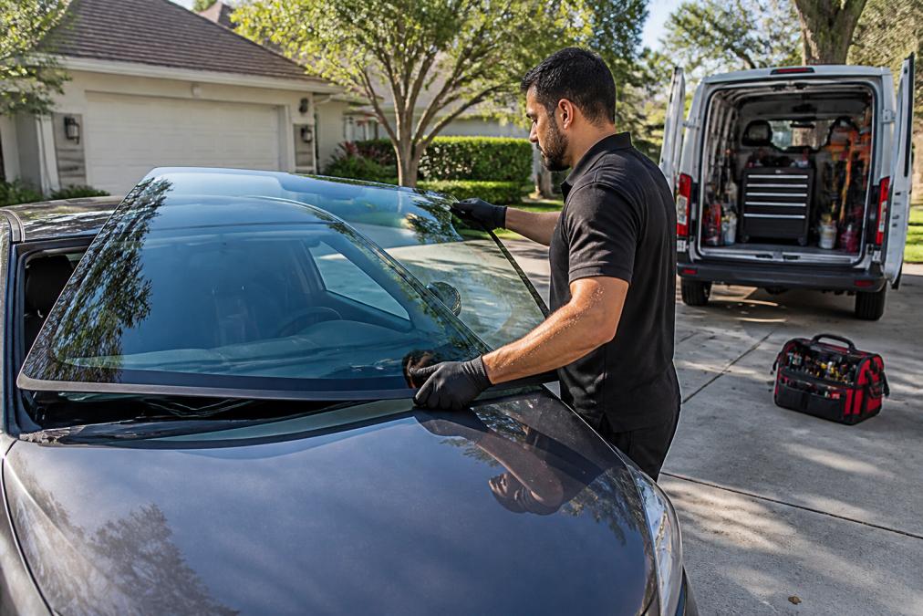 Auto glass technician in a branded uniform carefully lifting a new windshield into position on a sedan in a residential driveway, with a mobile service van and tool kit visible in the background under bright morning light