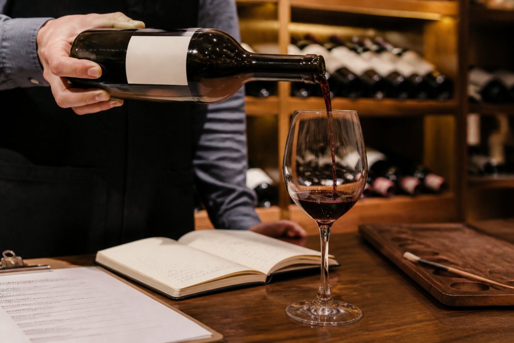 Close-up of a sommelier's hand pouring wine from a bottle into a wine glass, showing the standard 5-ounce fill line, with wine list and tasting notes visible in soft background lighting