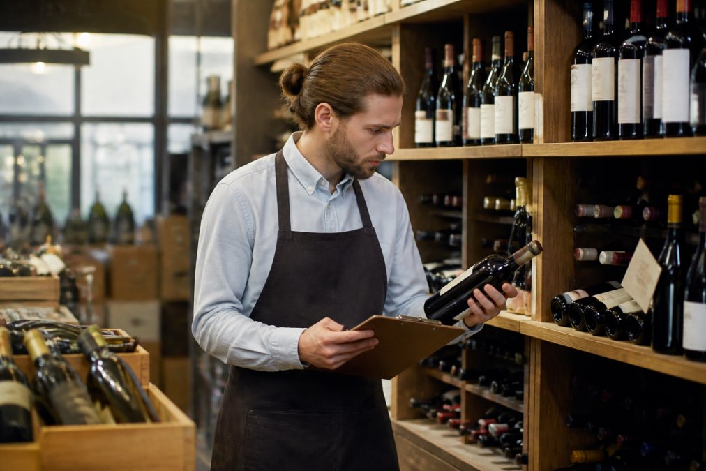 Sommelier in restaurant cellar reviewing wine inventory on tablet, bottles organised by region on wooden racks, warm lighting from vintage fixtures