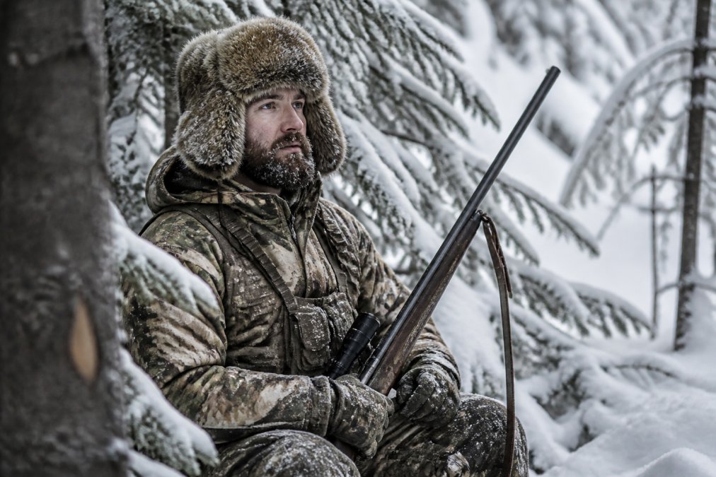 Hunter wearing fur hat and cold-weather gear in snowy forest, demonstrating proper fit and field functionality