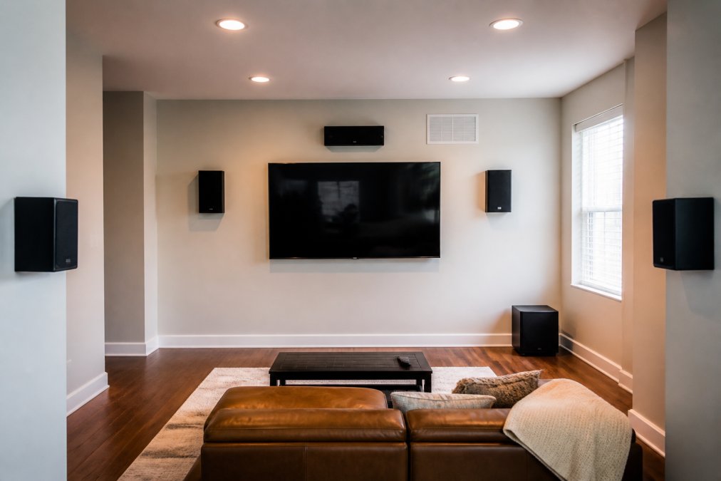Living room with a TV mounted on the wall, front left and right speakers positioned at ear level on either side of the screen angled inward, center channel speaker mounted above the TV, with a subwoofer visible in the corner and rear surround speakers mounted on side walls