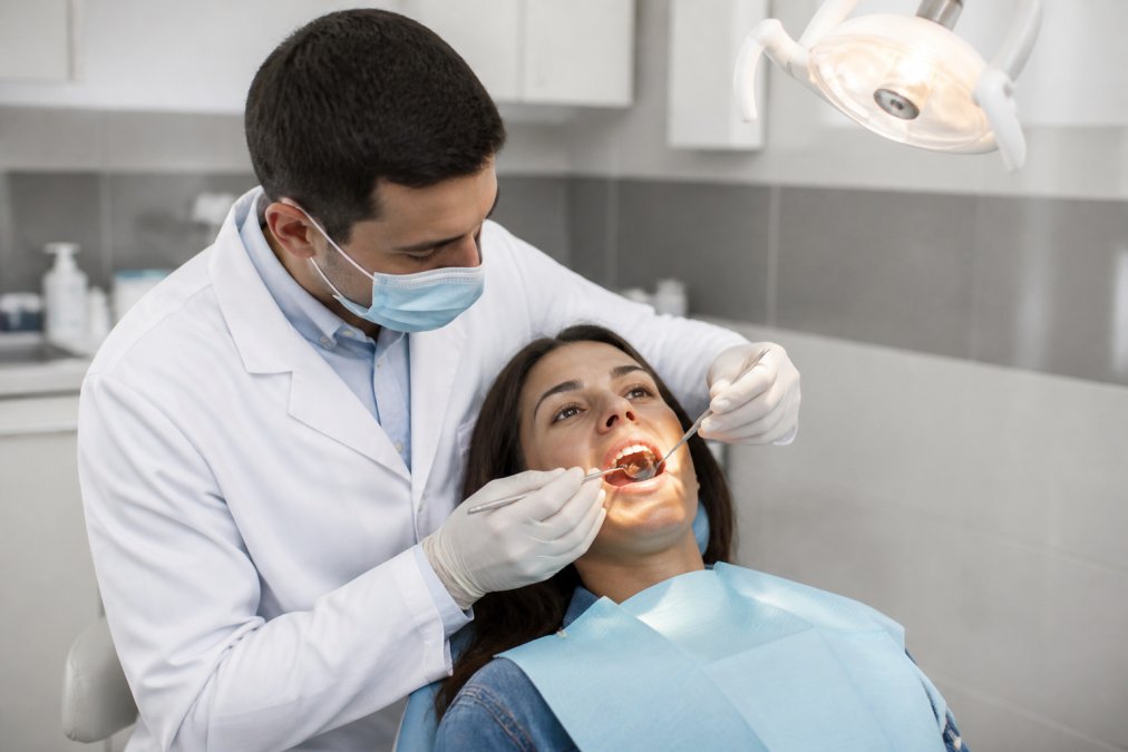 Dentist examining patient's mouth with overhead clinical light, using dental mirror and explorer tool during assessment