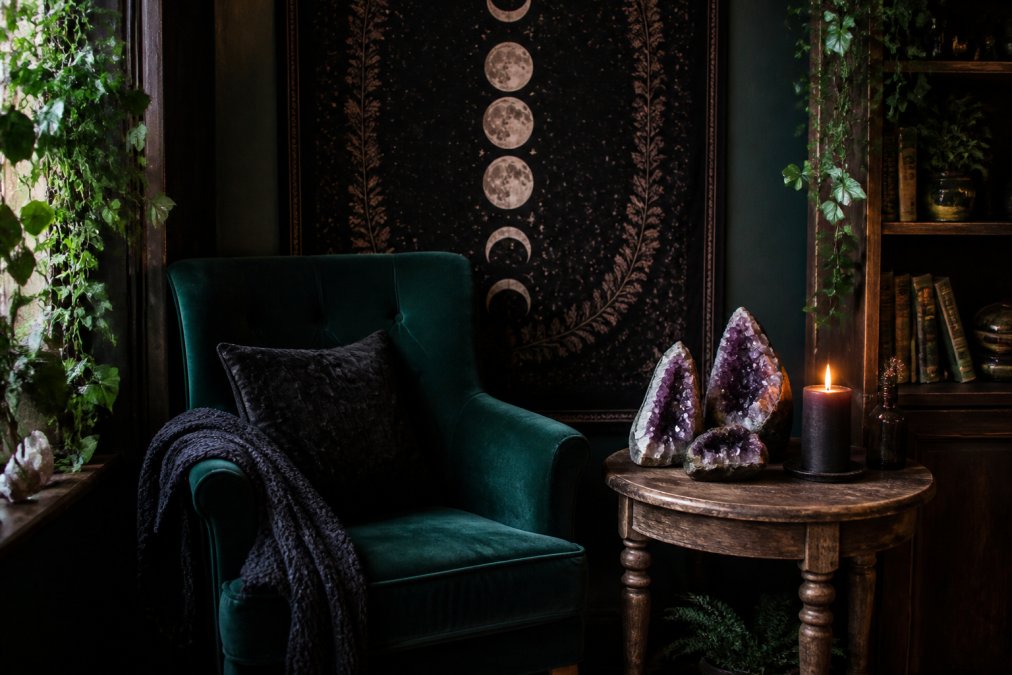 A moody corner of a living room styled with witchcore decor: a deep green velvet armchair, a rustic wooden side table holding a cluster of amethyst crystals and a lit pillar candle, and a large moon phase tapestry hanging on the wall behind it