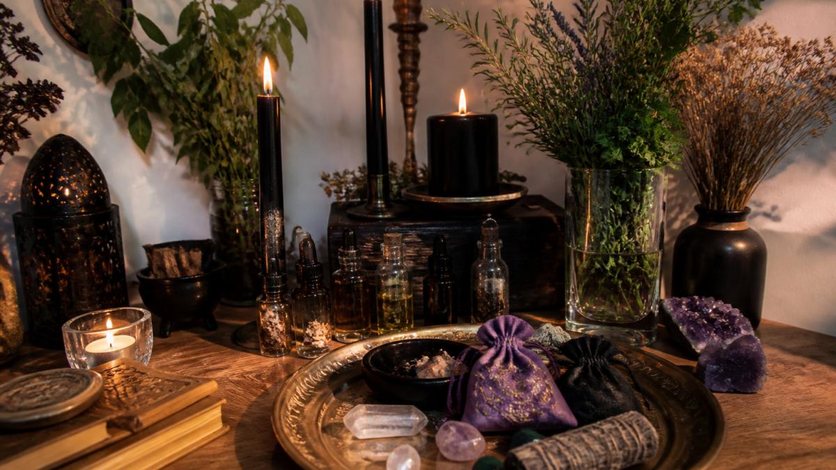 Neatly arranged home altar with lit spell candles, small bottles of ritual oil, fresh protective herbs in a glass vase, a ritual tray holding crystals and charm bags, and soft candlelight creating shadows on a wooden surface