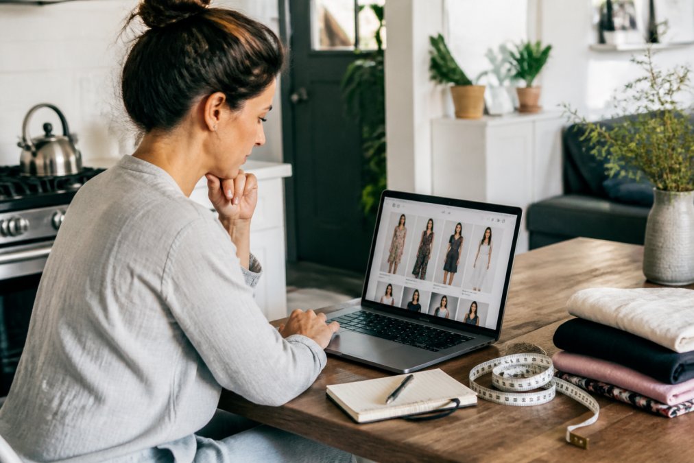 A woman in her thirties sitting at a sunlit kitchen table with a laptop open to an online clothing store, a measuring tape and a small notebook beside her, browsing dresses on the screen