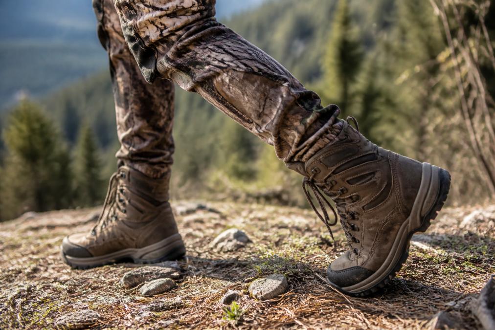 Close-up of woman's feet in properly fitted hunting boots on natural ground, showing correct boot fit with heel seated firmly, proper ankle support, and articulated knee movement during a walking stance in realistic outdoor lighting