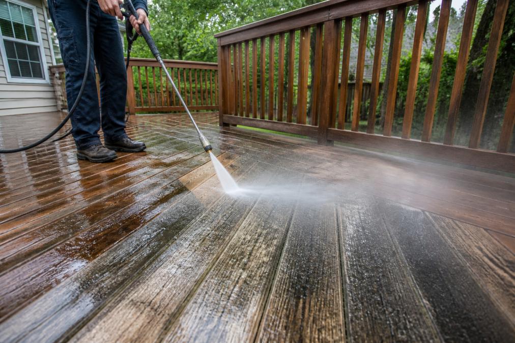 Professional demonstrating pressure washing technique on wooden deck surface, showing water spray pattern and mist on aged wood planks in natural daylight