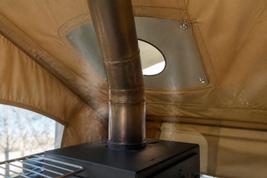 Close-up of a stove jack installed through canvas tent fabric, showing the metal ring, flue pipe connection, and fireproof mat underneath with visible heat shimmer