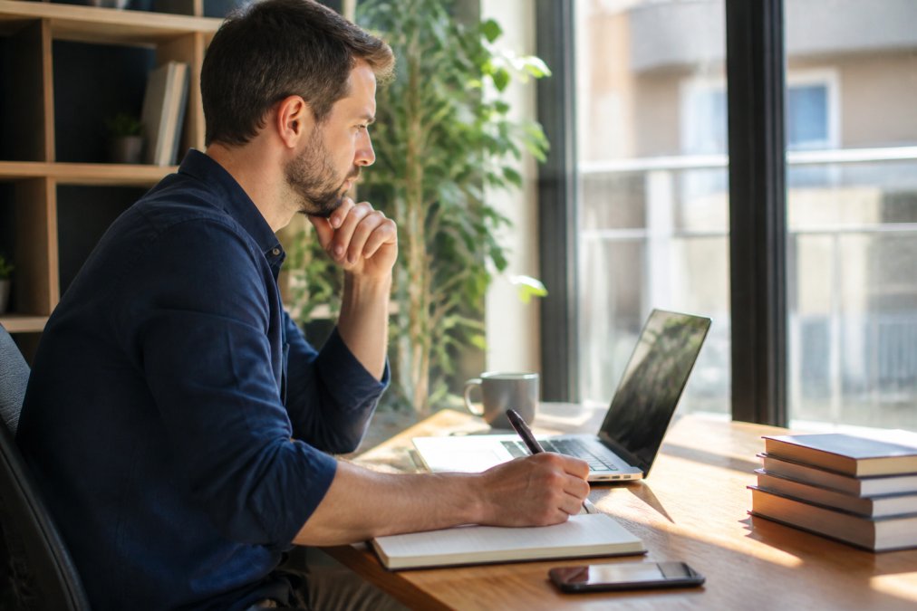 Unternehmer sitzt konzentriert an seinem Schreibtisch und überprüft sein Online-Shop-Dashboard auf einem Monitor, mit Notizen und Kaffee neben sich, natürliches Licht fällt durch große Fenster