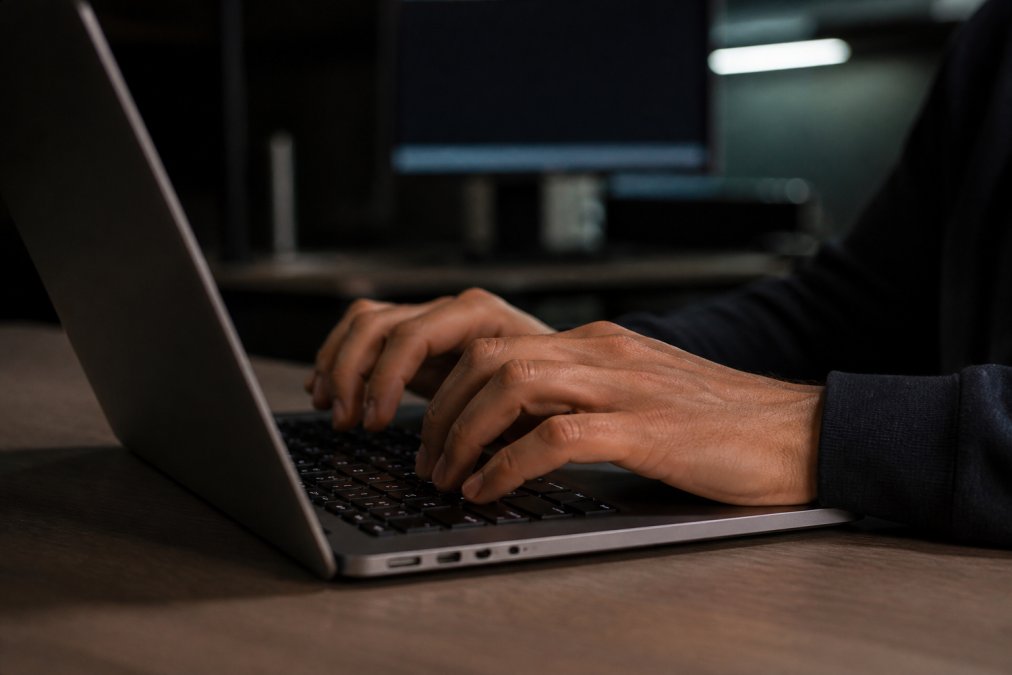 Close-up of hands typing on laptop keyboard with green security lock icon glowing on screen, dark office background with soft monitor lighting